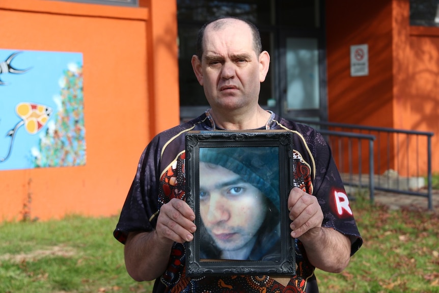 A sad man hold a framed photo of a young man wearing hoodie.
