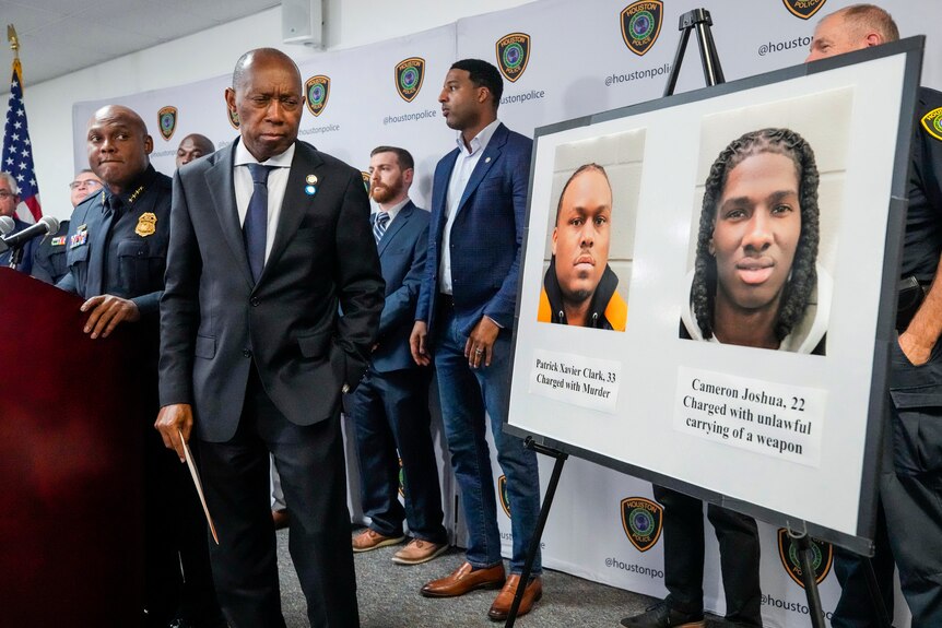 A black man in a suit stands at the front of a press conference next to a sign with two mjugshots on it. 