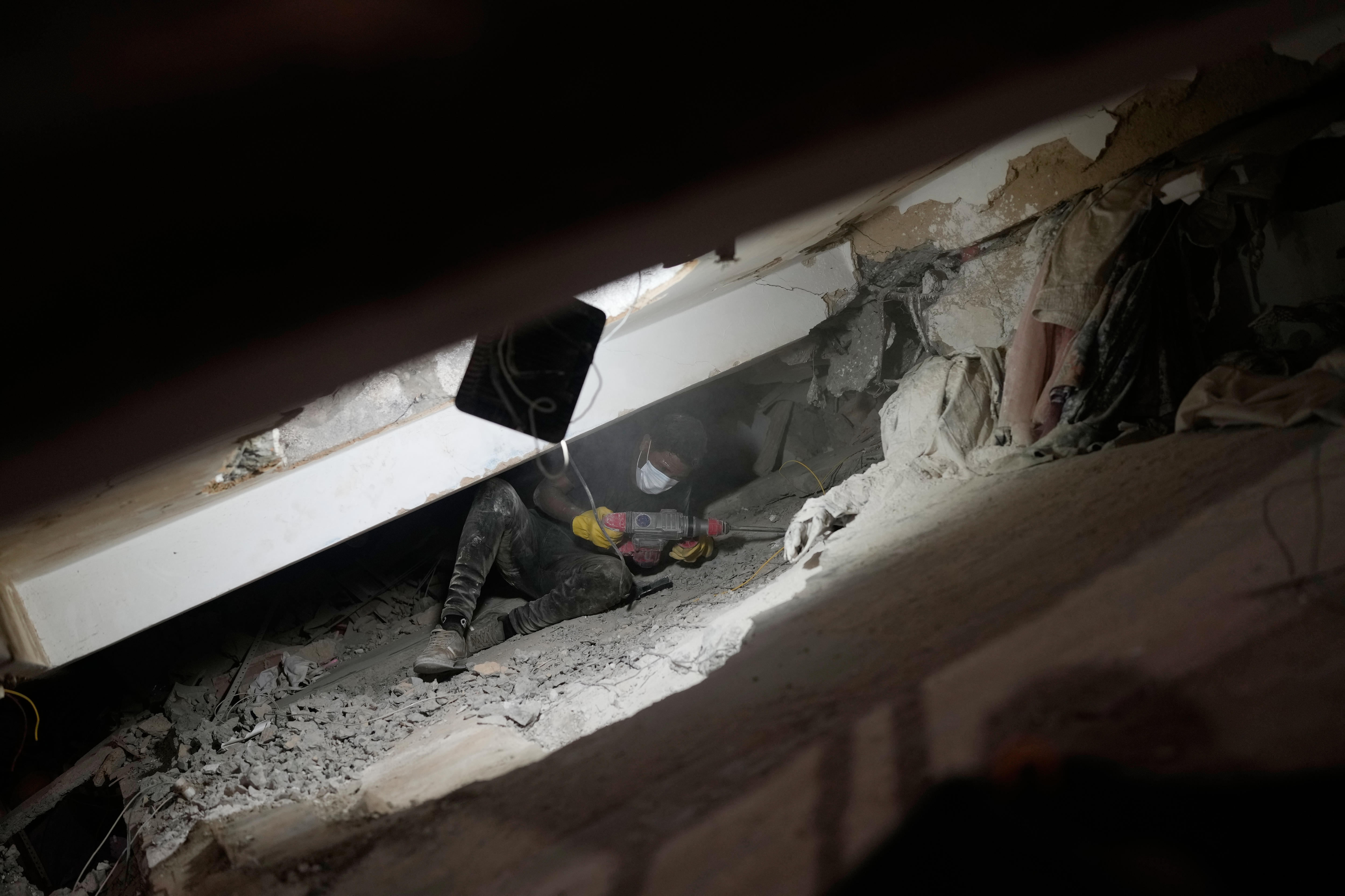 A man in dark clothing and a face mask, lying under a slab of concrete in a sliver of light next to a machine jack.