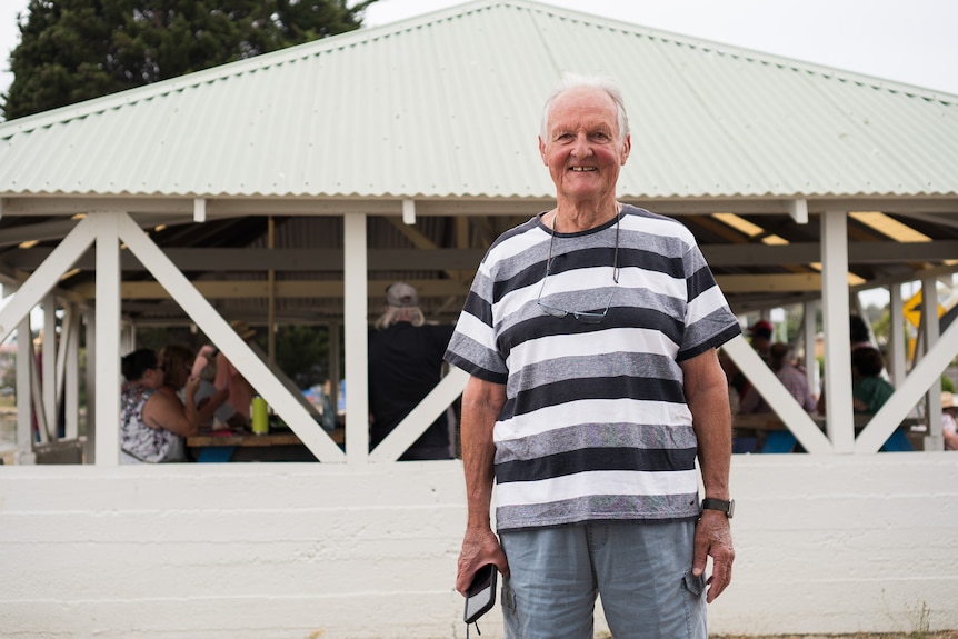 A man with white hair and a black, white and grey striped top smiles in front of a barbecue shelter.