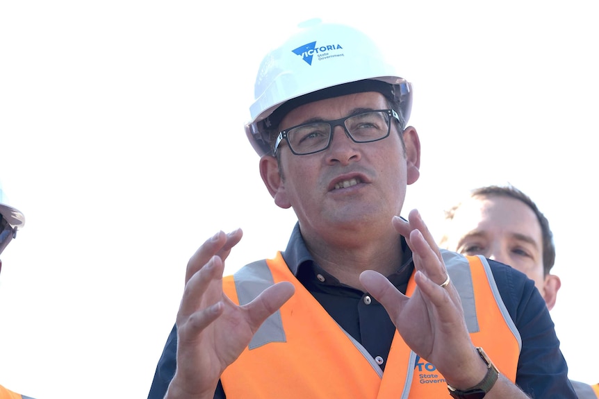 Daniel Andrews, wearing a hard hat and hi-vis vest, speaks at a press conference.