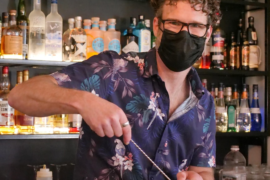 A man stands in front of a wall of alcohol mixing a cocktail and wearing a mask.