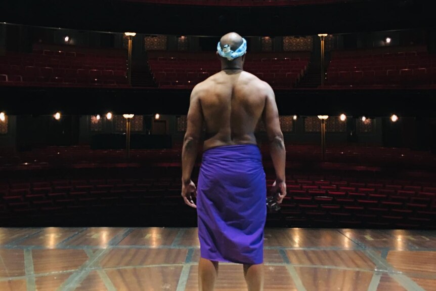 A shirtless, muscular Torres Strait Islander man wearing a cloth wrap around his waist on stage at a theatre with back to camera