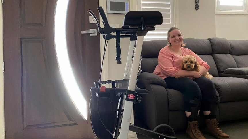 Jess sits smiling on a couch with her dog Riley, next to a gutter frame mobility aid. Sun shines through glass in the door.
