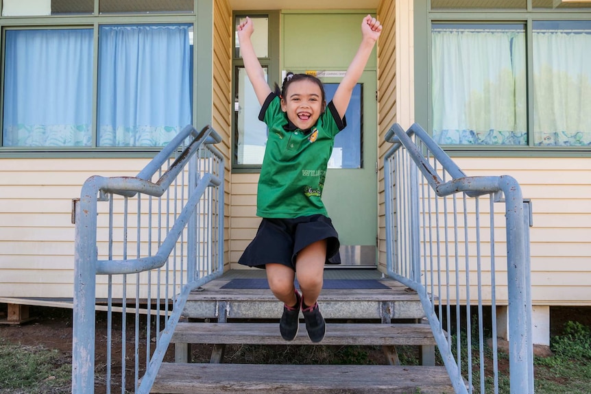 A young girl jumps off a stair with her arms and legs in the air. She is smiling widely.