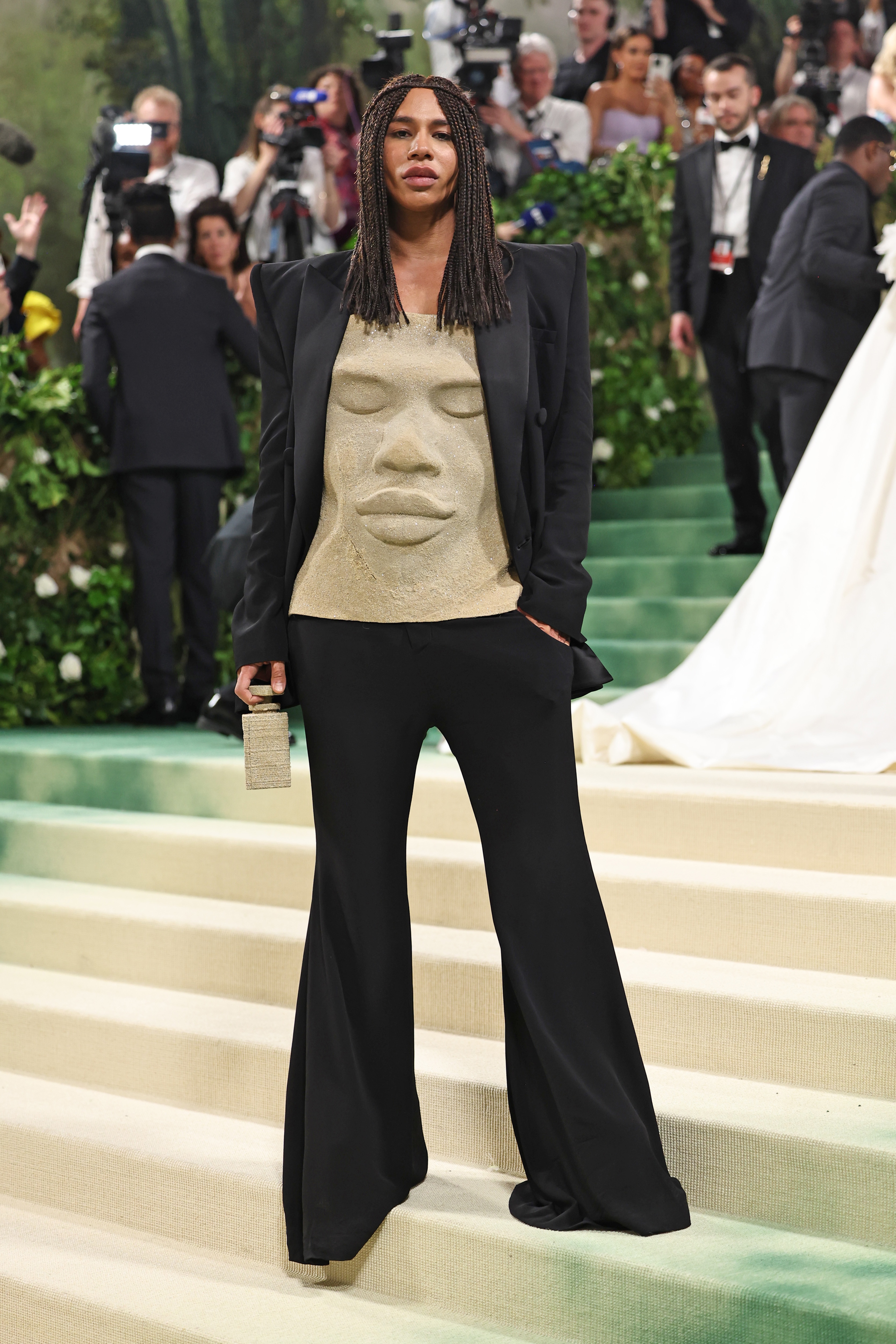 A black man with braids on the Met Gala steps in black pants and blazer, with a shirt made of sand in the shape of his face.