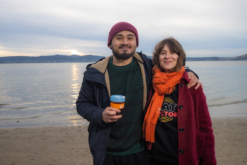 A young couple in warm jackets stand on a beach and smile