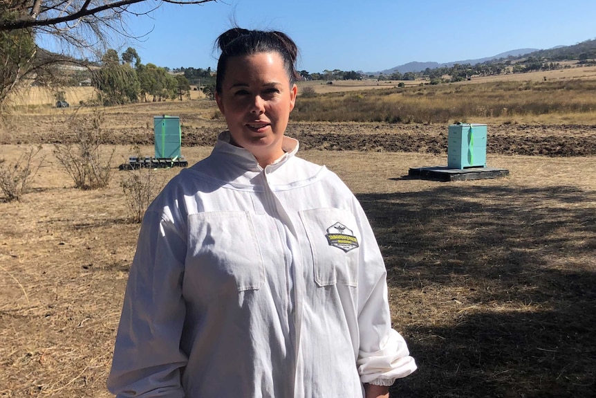 A woman in a bee keepers protective suit stands in front of three hives