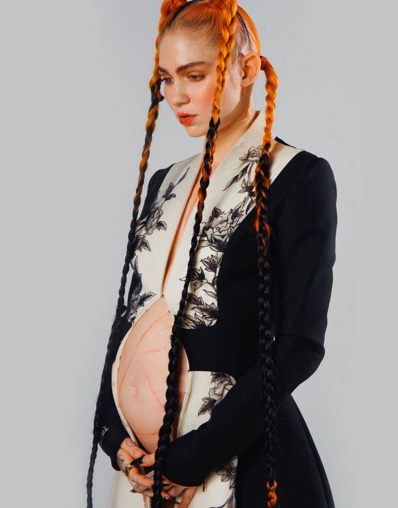 Grimes holds her pregnant belly, which exposed. She wears a long black coat and long orange plaits in her hair. 