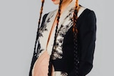Grimes holds her pregnant belly, which exposed. She wears a long black coat and long orange plaits in her hair. 