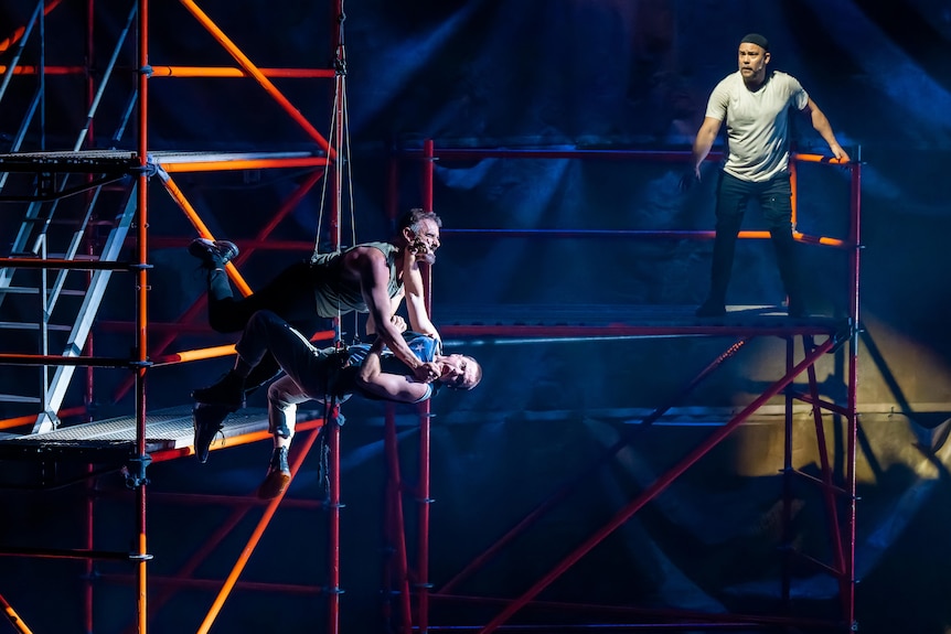 Two harnessed men, one Middle Eastern, one white, tousle as they fall from scaffolding on stage. An Aboriginal man watches.