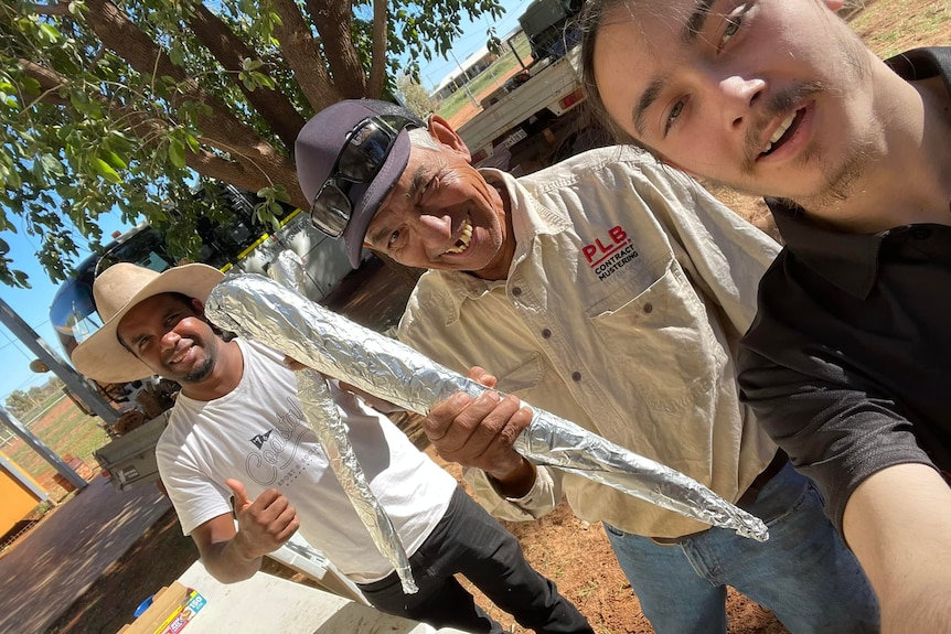 A male teenager takes a selfie with two men, one holding a kangaroo tail wrapped in alfoil, the other doing a thumbs up.