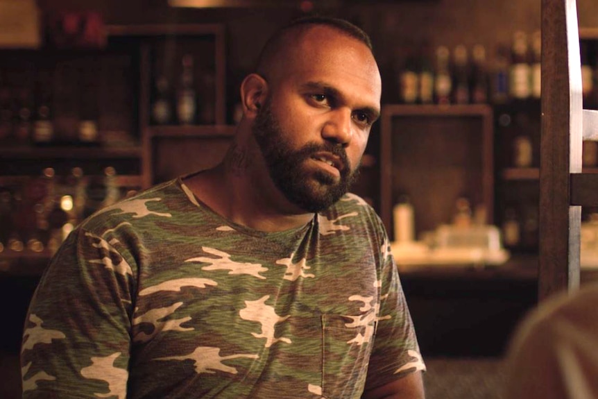 A young man with a camo t-shirt sits in front of a bar.