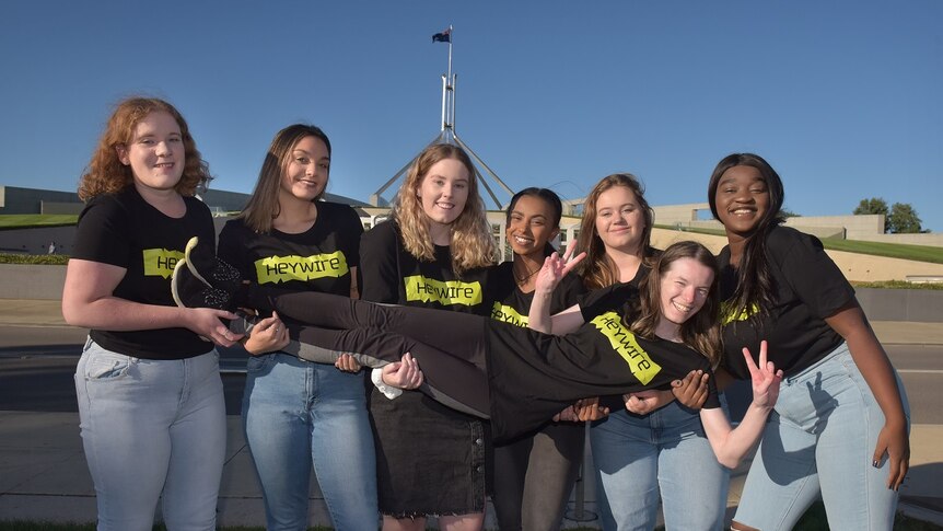 Six young women in Heywire shirts are hold up a seventh woman in front of Parliament House.