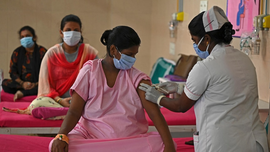 A woman sits cross-legged on a hospital bed while a healthcare worker injects a vaccine into her arm. 