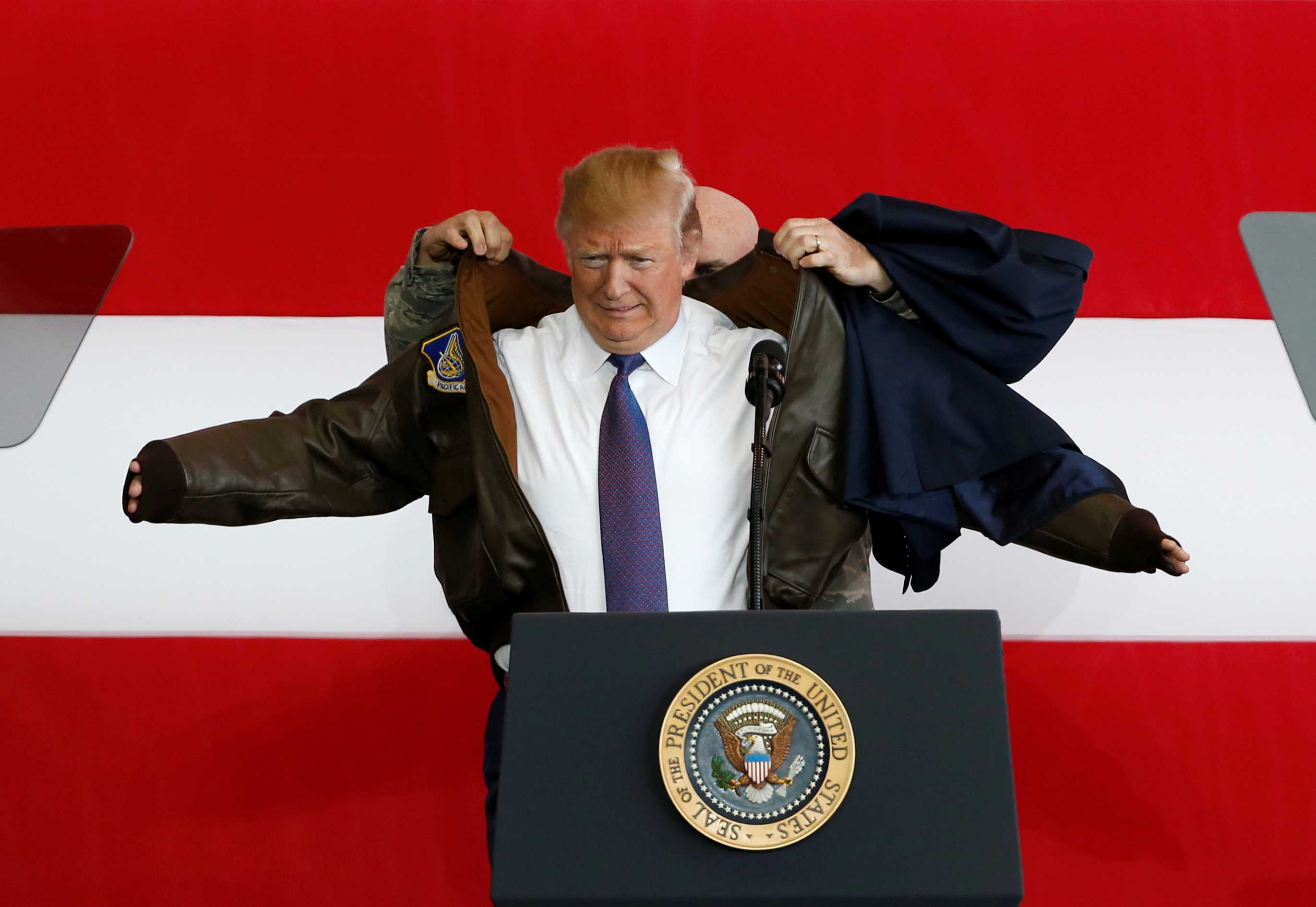 A man helps another man put on a flight jacket in front of a large American flag.