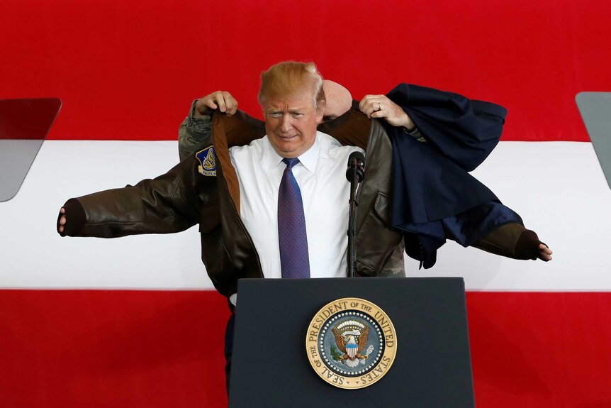 A man helps another man put on a flight jacket in front of a large American flag.