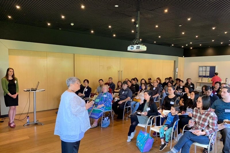 A woman speaks to an audience of people, seated in rows.