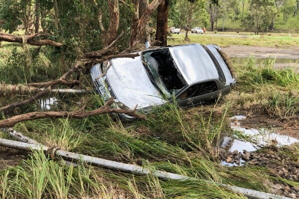A grey sedan (Toyota Camry) washed off the road and onto trees, the car is on it's side, windows smashed surrounded by debris