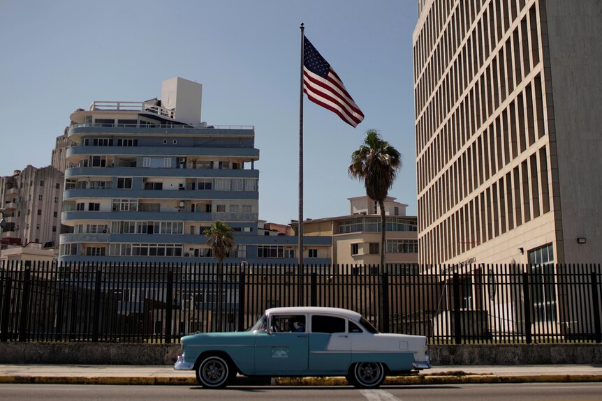 A teal and white car drives by the US Embassy in Havana, Cuba with a large American flag on a flagpole. 