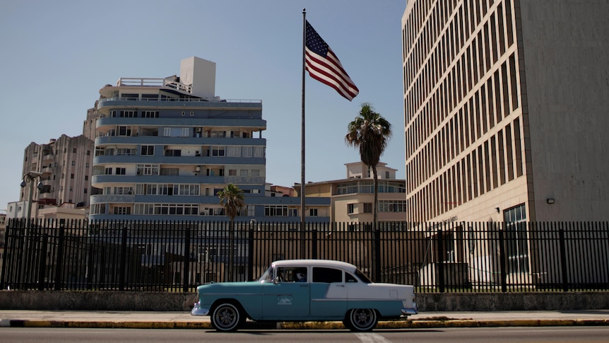 A teal and white car drives by the US Embassy in Havana, Cuba with a large American flag on a flagpole. 