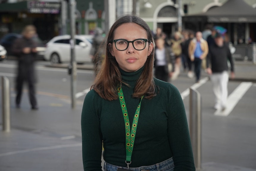 A young white woman with long brown hair standing int he street. She's wearing a bright green lanyard with sunflowers on it