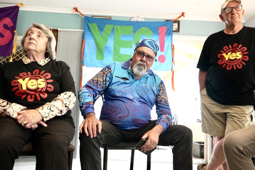 Wally sits starring down at the camera between two people in Yes t-shirts