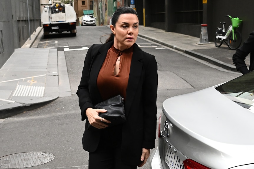 Vanessa Amorosi wears a black jacket and pants and brown top and holds a black bag as she walks behind a white car.