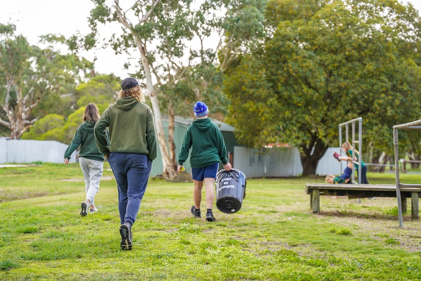 A school student walks through a grassed playground carrying a black waste bin.