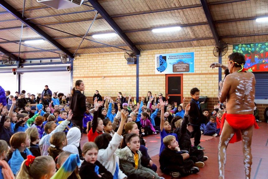 An Aboriginal man dressed in traditional clothes and body paint performing at a school.