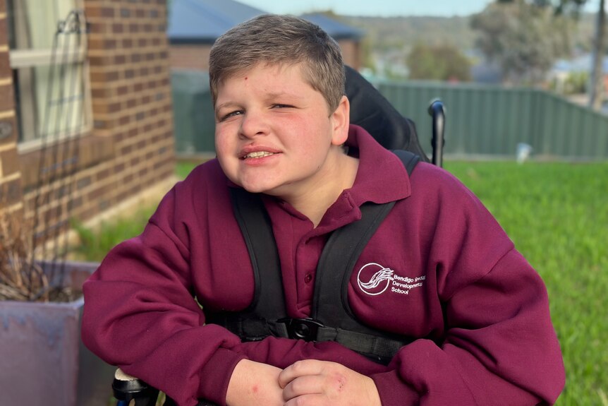 A 12 year old boy sitting in a wheelchair wearing a maroon school jumper.