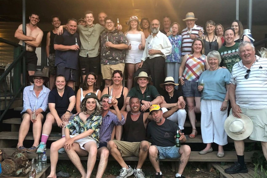 About 30 people are sitting and standing on a porch looking at the camera with drinks in their hand.