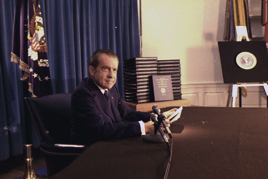 Richard Nixon sits at a desk looking into the camera