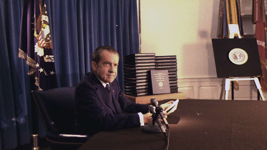 Richard Nixon sits at a desk looking into the camera