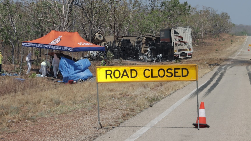 Road closed sign on the side of road with crashed truck and car behind it in the bush