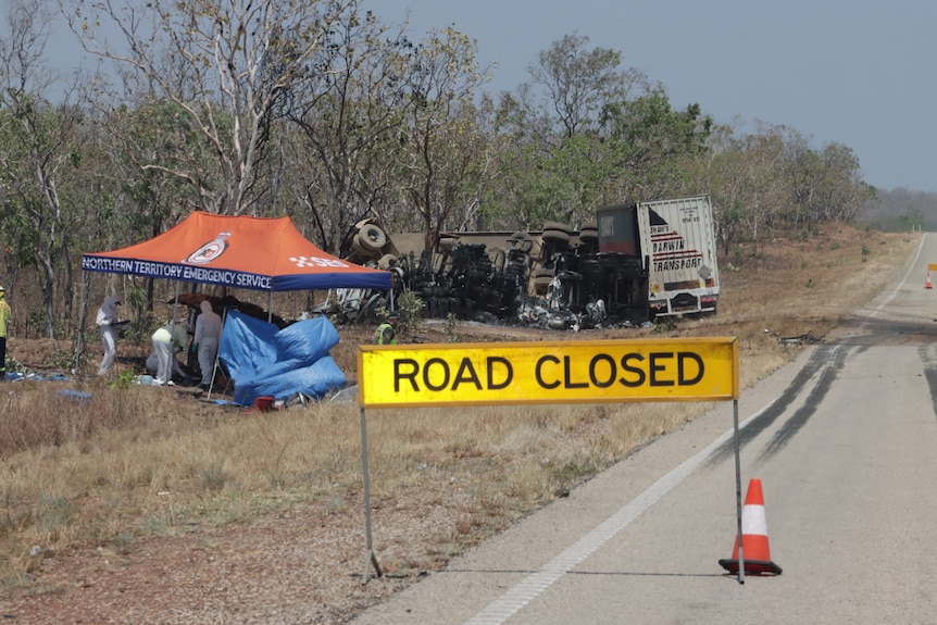 Road closed sign on the side of road with crashed truck and car behind it in the bush