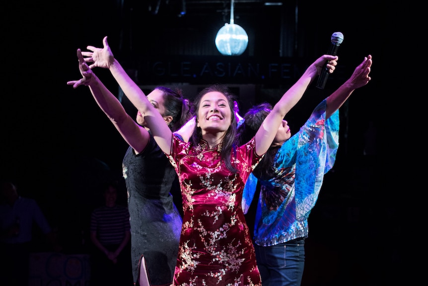 Three Asian Australian woman stand back-to-back on a podium under a disco ball with arms outstretched and beaming smiles.