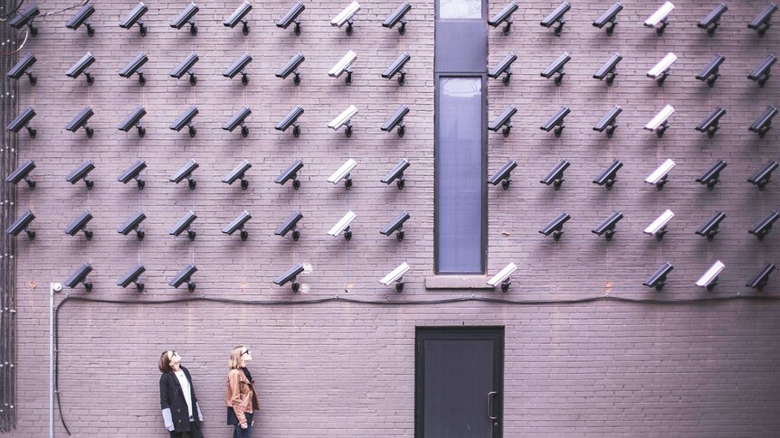 Two women standing beside a wall that is covered in identical rows of security cameras.