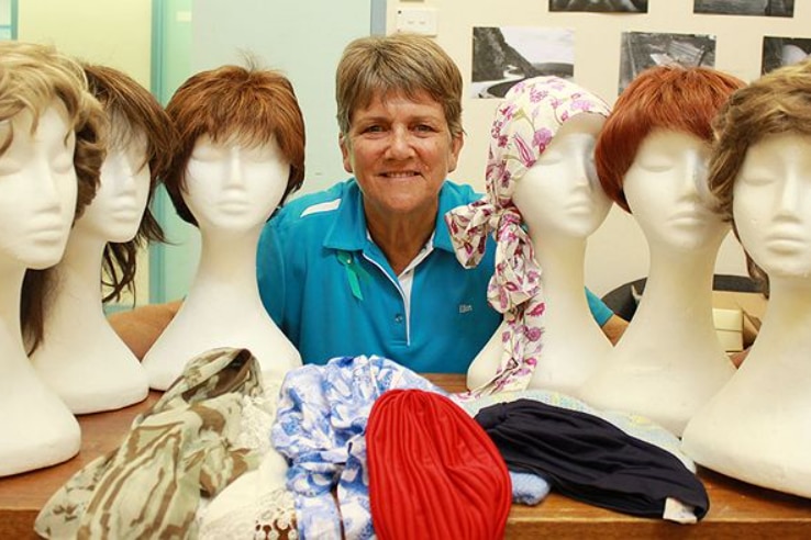 A woman has short hair, a blue shirt and smiles. She is surrounded by wigs on mannequins, and colourful turbans