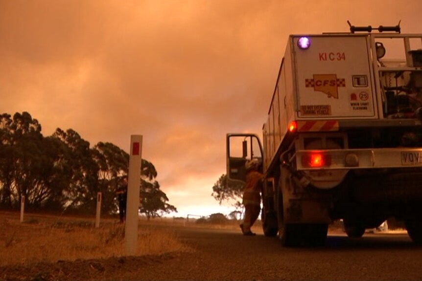 A firefighter getting into a fire truck with an orange glow behind trees
