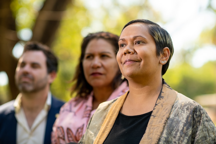 A woman smiles seriously at the camera. Behind her are a man and a woman.