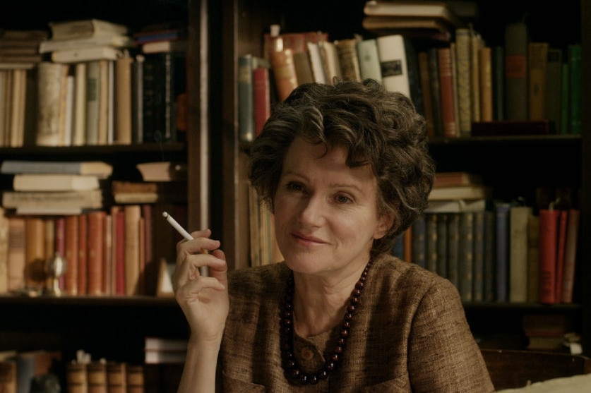 A woman with 1960s brown permed hairdo in brown top and bead necklace sits at desk with cigarette in front of large bookcase.