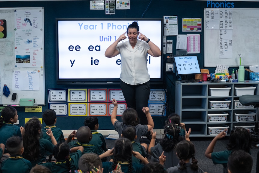 A teacher stands in front of a class of year 1 students, pointing at her head, in front of a slide show about phonics.