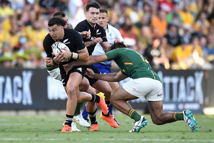 An All Blacks player holds the ball as two Springboks opponents try to tackle him.