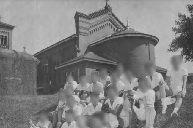 Black-and-white photograph of men in the 1960s wearing cricket uniforms, watching a game from a hillside.