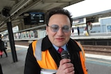 A man in a high-vis vest on a train platform holds a microphoneclose to his face, his eyes closed in concentration.