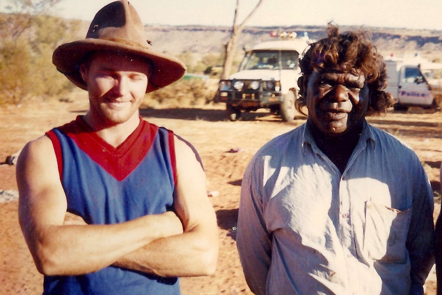 A man in a worn brown hat and blue and red footy jumper smiles with his arms crossed next to a smiling man in a white shirt.