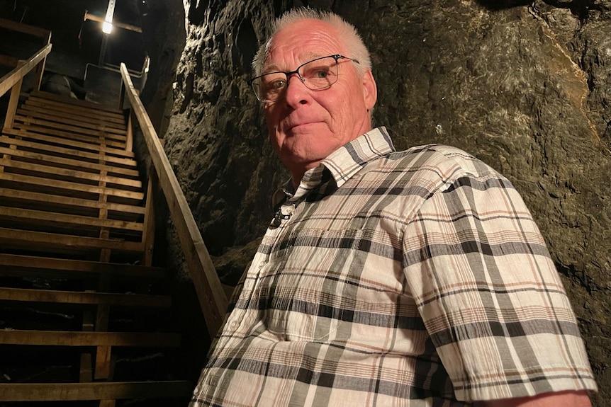 An older man wearing glasses stands next to a steep staircase inside an underground bunker.