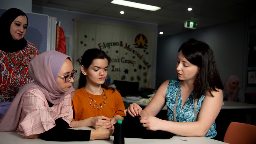 Gina Bajeel teaches refugee women dress making skills at a table with fabric in hand as they look on.
