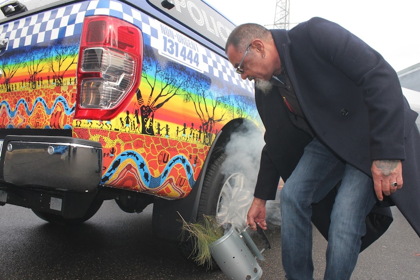 A man holds a tin of smoking leaves near a colourful police vehicle